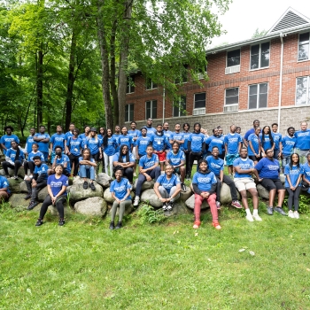 A group of people wearing blue shirts, adults, gather for a group picture. They're standing in grass area with tress in the background.