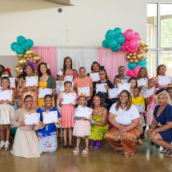 A group of people, including adults and children, posing for photo. A pink and white background with blue and pink balloons decorations.