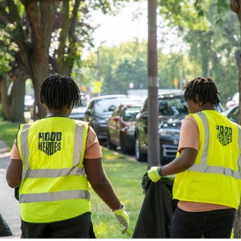 Two people wearing yellow emergency vests with Hood Heroes written on the back of them, holding a black garbage bag cleaning garbage off the ground.
