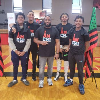 A group of young men smiling to the camera wearing black shirts with I AM CHI written on them. An African American flag stands on the side of the group.