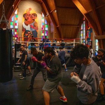 A group of boxers practicing a boxing stance with a boxing ring in the background. They're in a large room with a painted mural of two boxers.