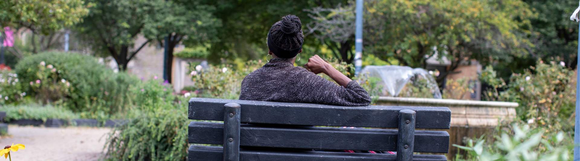 A person sits on a wooden park bench, wearing a dark sweater and a headscarf, facing away. The background features a garden with trees and flowers, and a stone fountain is visible under overcast skies.