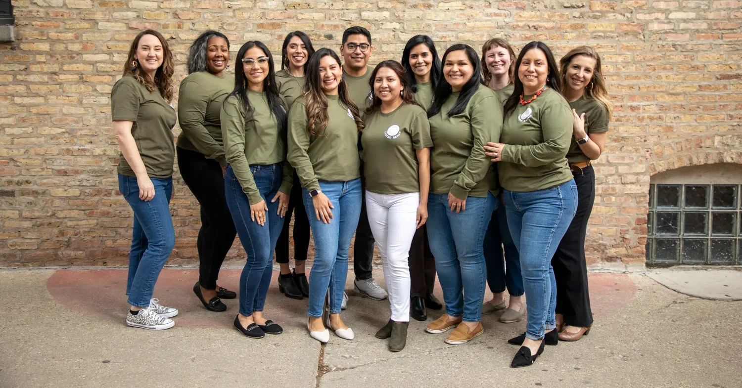 A group photo of the men and women smiling towards the camera and wearing matching Centro Sanar shirts. The background features a light brick wall.