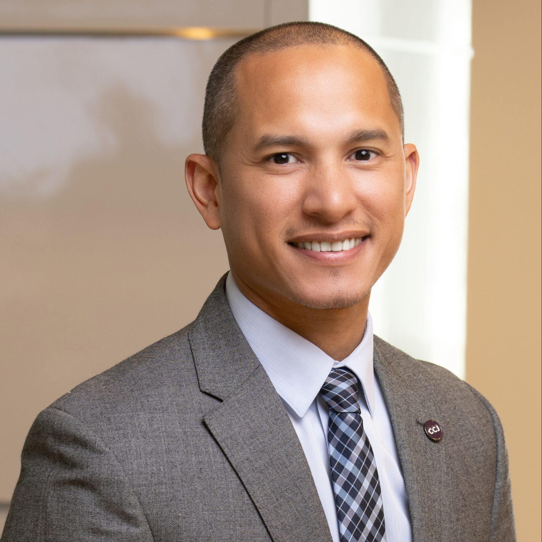 A man wearing a gray business suit, smiling at the camera. The background is a light tan color.