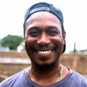 A man wearing a hat backwards and a purple shirt, smiling at the camera. The background is a background of a city backgrounds.