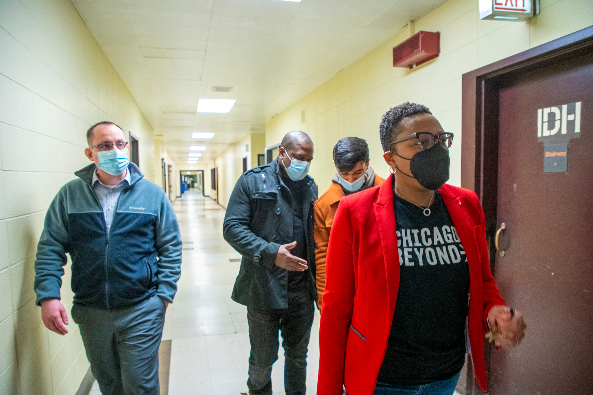 A group of four people, wearing face masks, walk down a hallway. The person in front wears a red blazer and a Chicago Beyond t-shirt. The hallway has beige walls, a tiled floor, and an EXIT sign above a door.