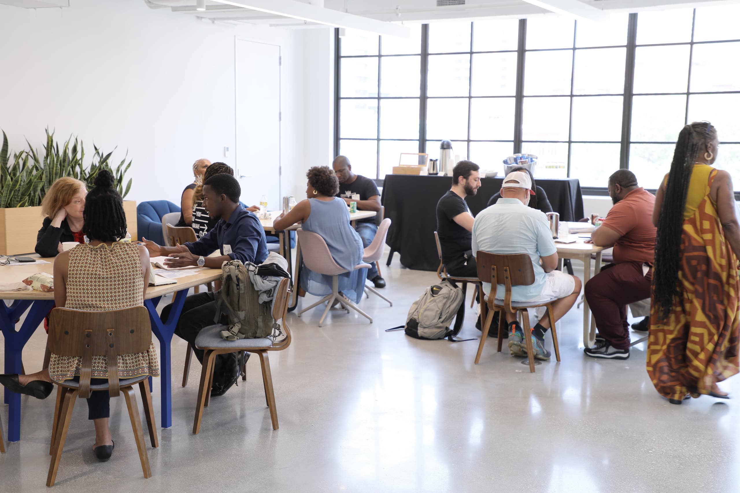 A group of people sitting down at three tables in a room with white walls and windows in the day time.