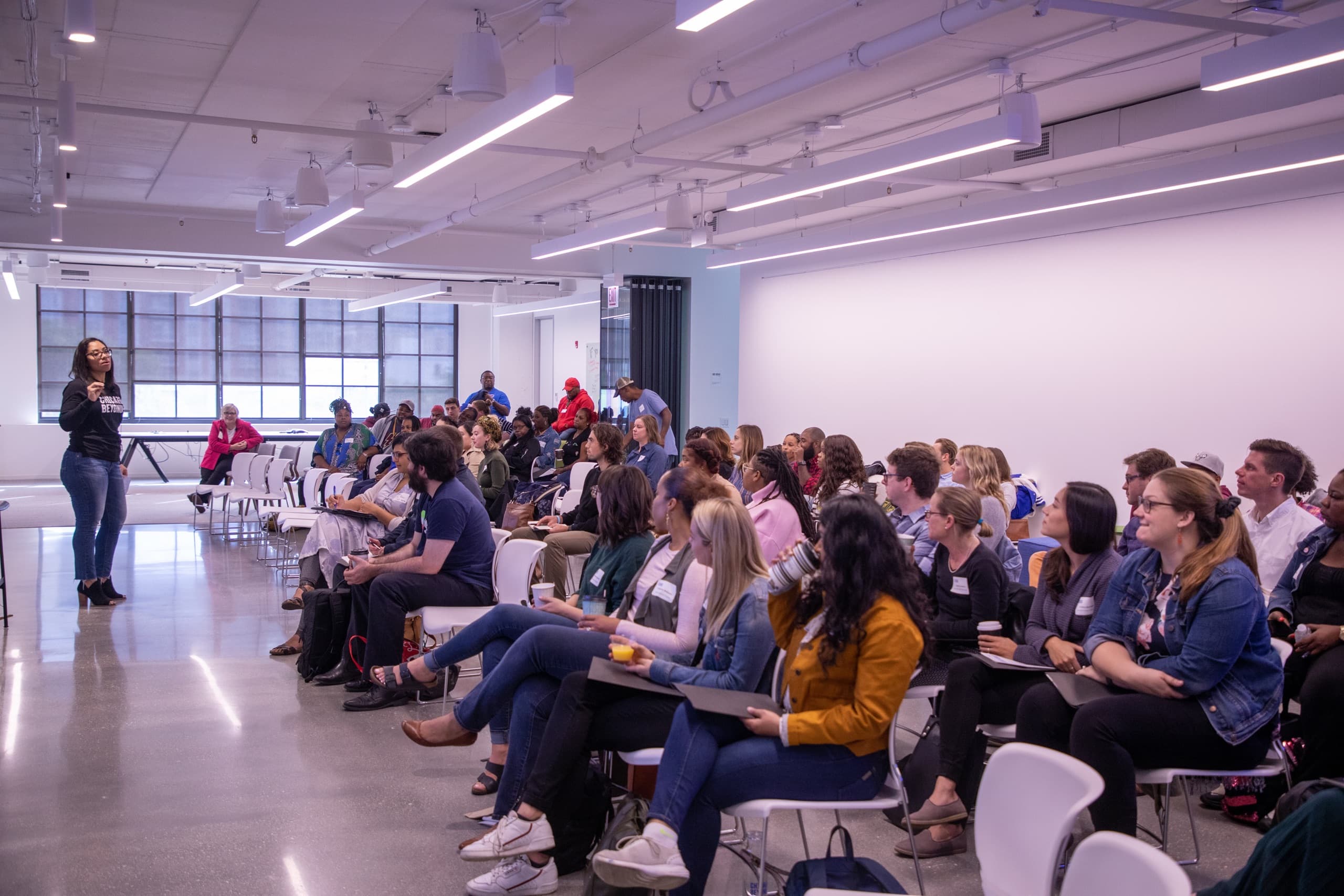 A group of people sitting down in rows, they are looking towards a women speaking towards them. The background has windows in the distance and a white wall behind the audience chairs.