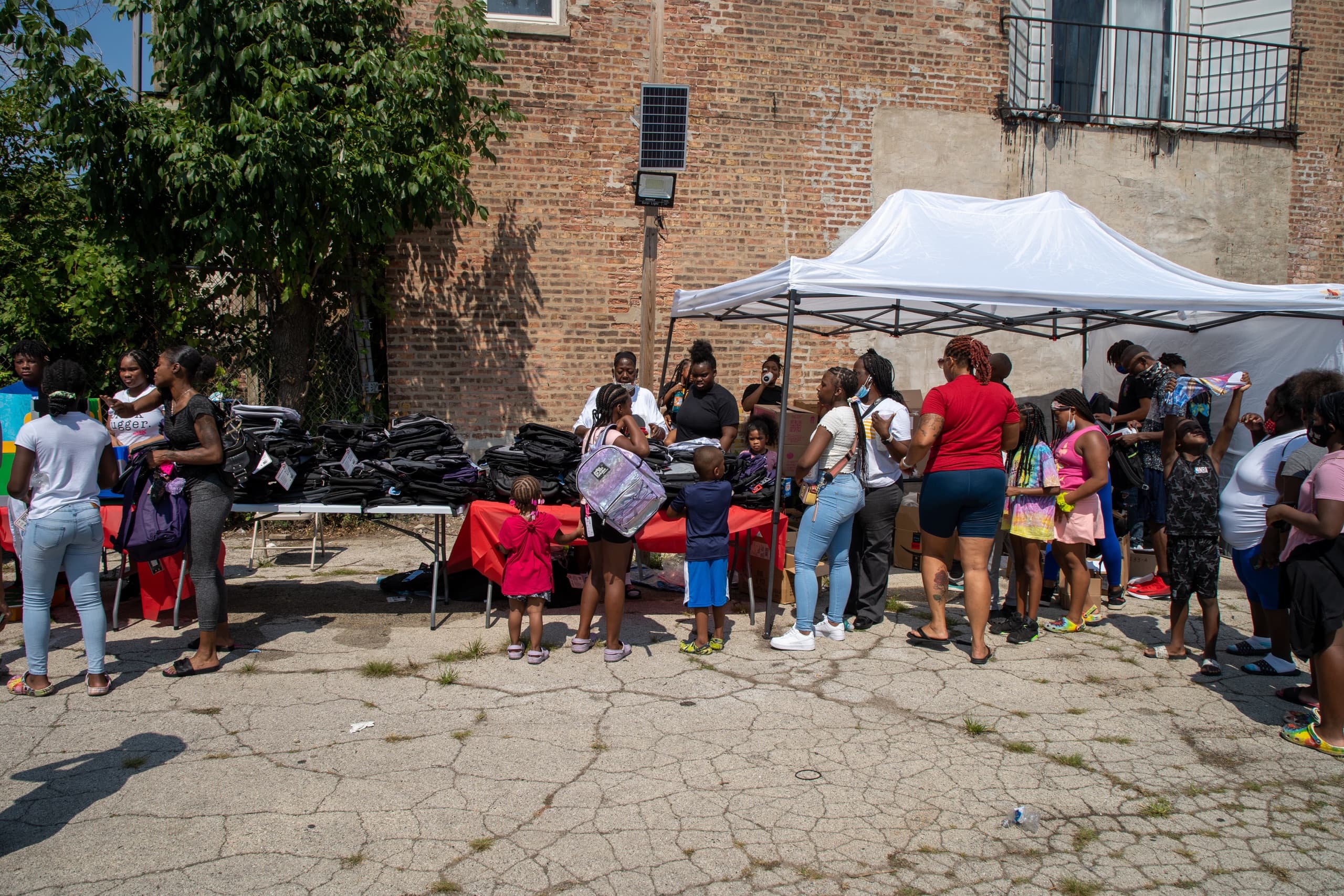 A group of people, including adults and children, gather around a tent set up outdoors. They are receiving backpacks and school supplies on a sunny day. A brick building and trees are visible in the background.