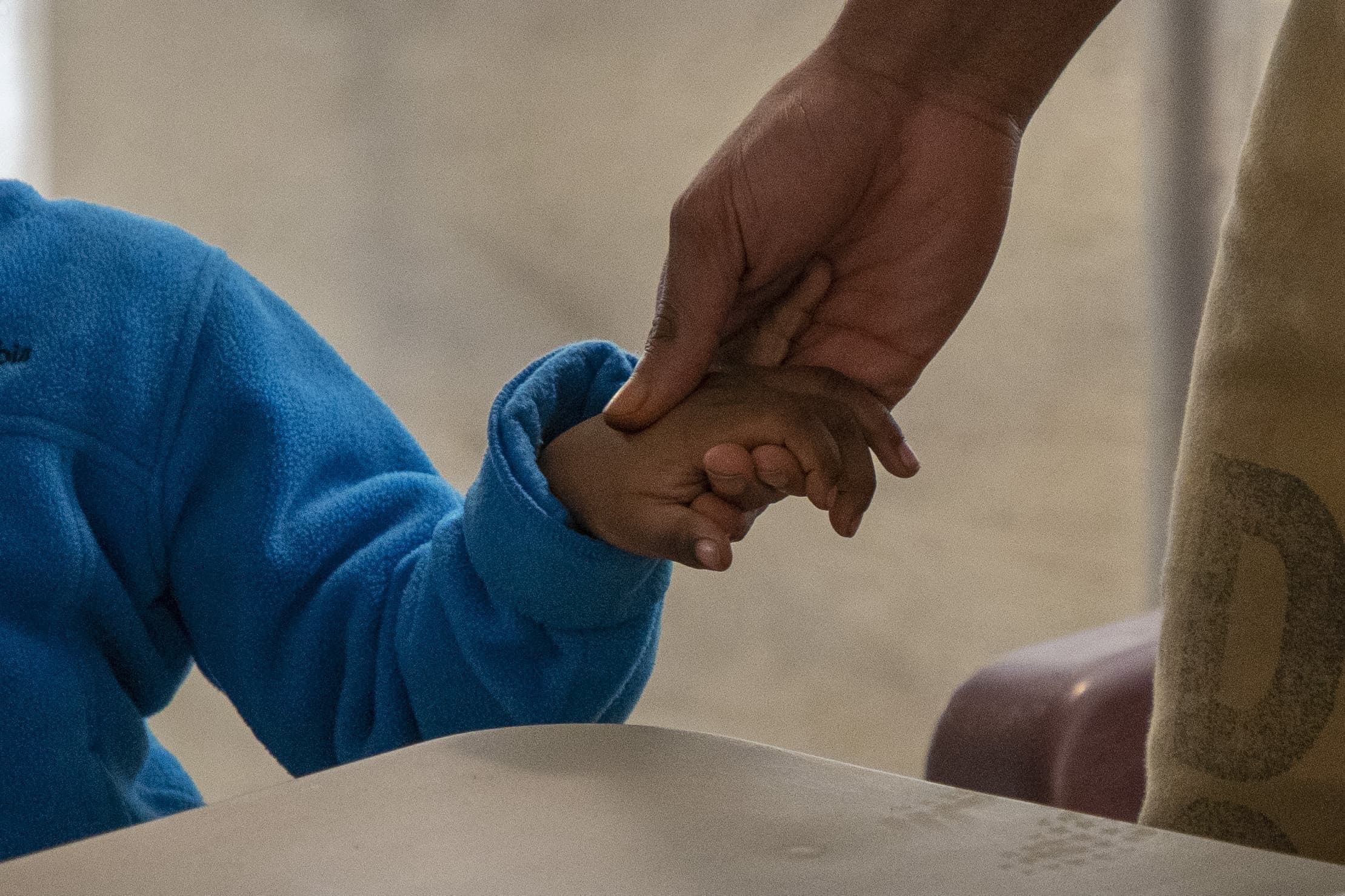 An adult hand gently holds a childs hand over a table. The child is wearing a blue sweater. The background is blurred.