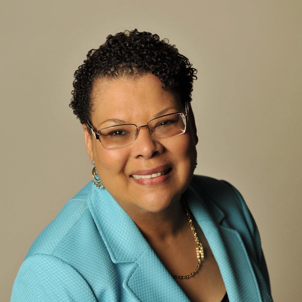 A woman wearing glasses and a light blue business suit, smiling at the camera. The background is a light tan color.