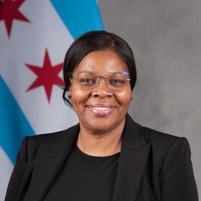 A woman wearing a black shirt and suit blazer smiling with a chicago flag behind her on a gray backdrop.