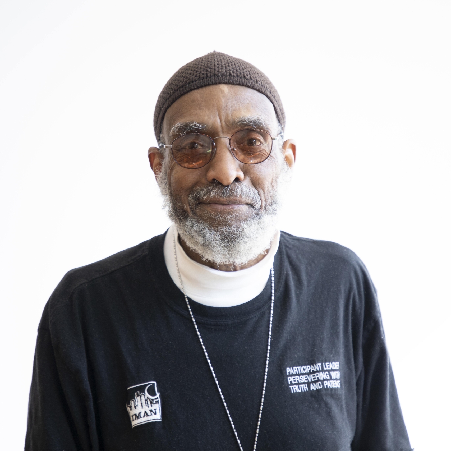 Person wearing glasses and a blue long sleeve shirt, smiling at the camera. On the shirt reads IMAN. The background is a light white color.