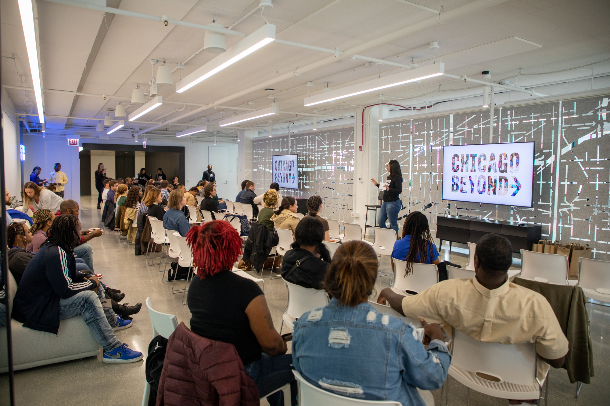 A group of people is seated in a bright, modern conference room, watching a presentation. A speaker stands at the front near a large screen displaying Chicago Beyond. Attendees are facing forward, engaging with the presentation.
