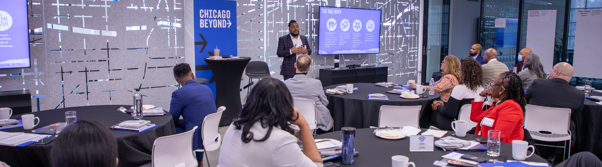 A man speaking to a group of people sitting at tables while he has graphics on display to the left and right of him. A Chicago Beyond sign is standing to the left side of the man. The room has a gray wall.