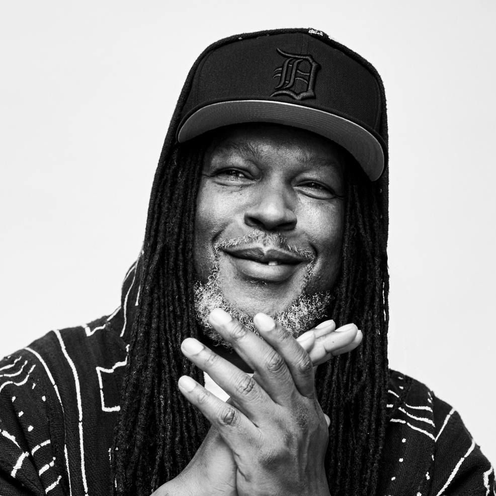 A man smiling at the camera with his hands clasped together wearing a hat with a letter D on it with a white background.