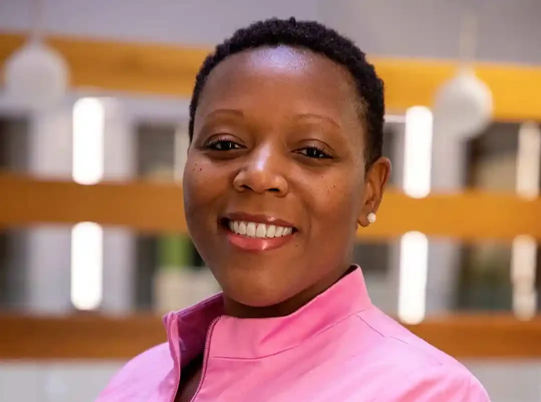 A woman wearing a pink jacket smiles at the camera. The background features blurred horizontal wooden slats and circular light fixtures.