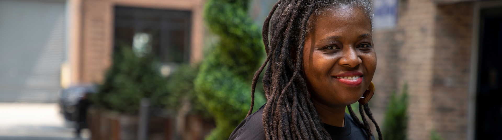 A woman with long dreadlocks smiles while looking into the camera. The background is filled with green shrubs.