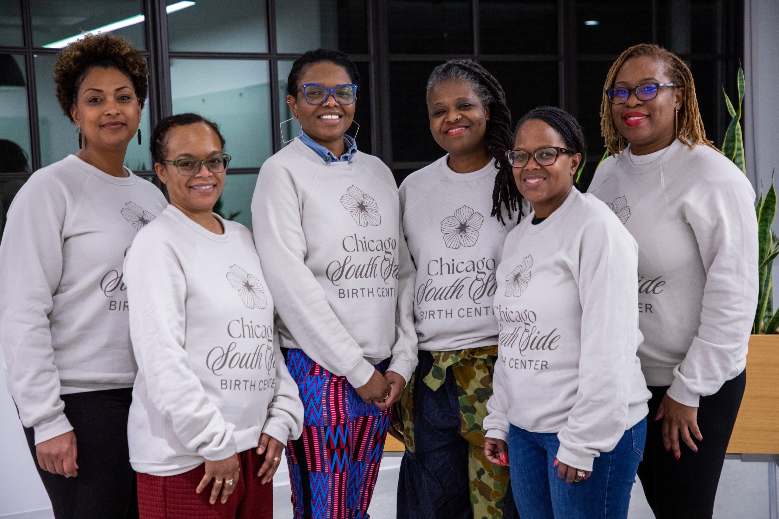 Six people standing indoors, smiling and wearing matching Chicago South Side Birth Center sweatshirts. They are positioned in two rows, with three in the back and three in the front. A plant and large windows are in the background.