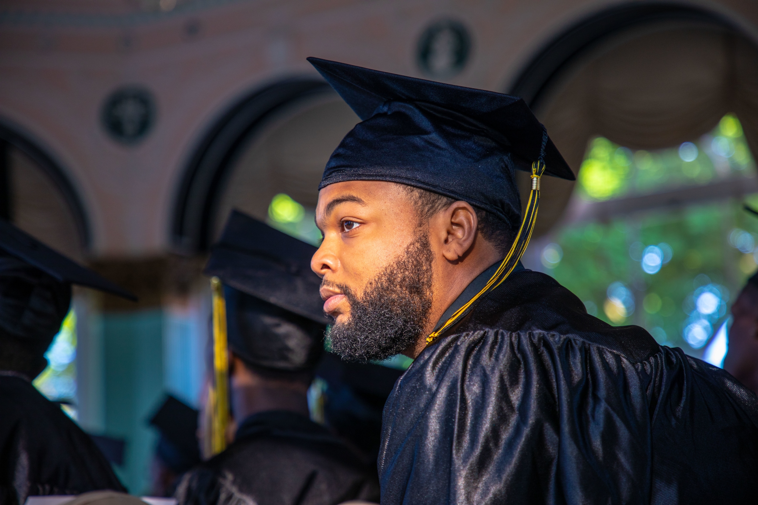 A graduate in a black cap and gown, with a yellow tassel, looks to the side during a ceremony. The background shows arched windows and blurred greenery outside, suggesting a formal event setting.