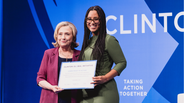 Two women stand on stage, smiling for a photo. One holds a certificate labeled Clinton Global Initiative Commitment to Action. The background features the words Clinton Global Initiative and Taking Action Together.