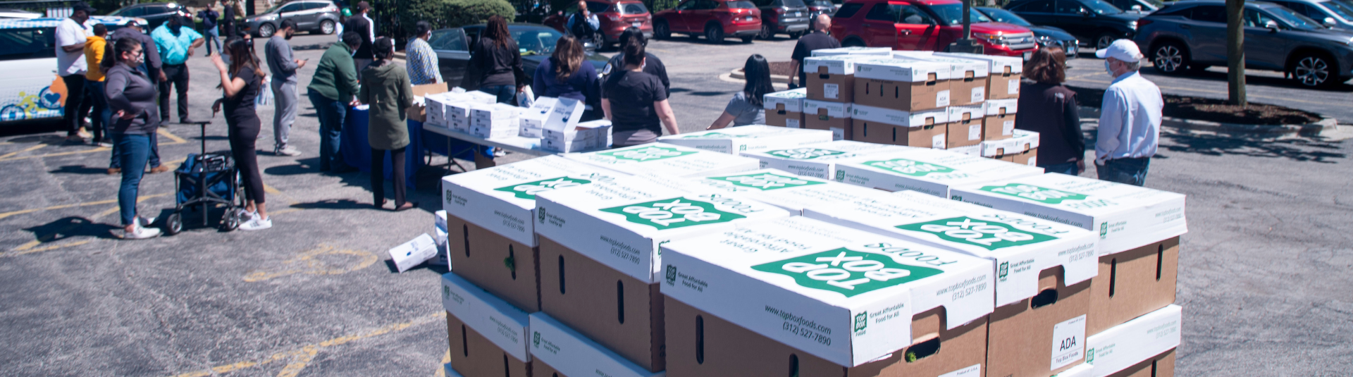 People gather outdoors near tables and stacks of boxes labeled Food Box. Cars are parked in the background, suggesting a community distribution event in a parking lot.