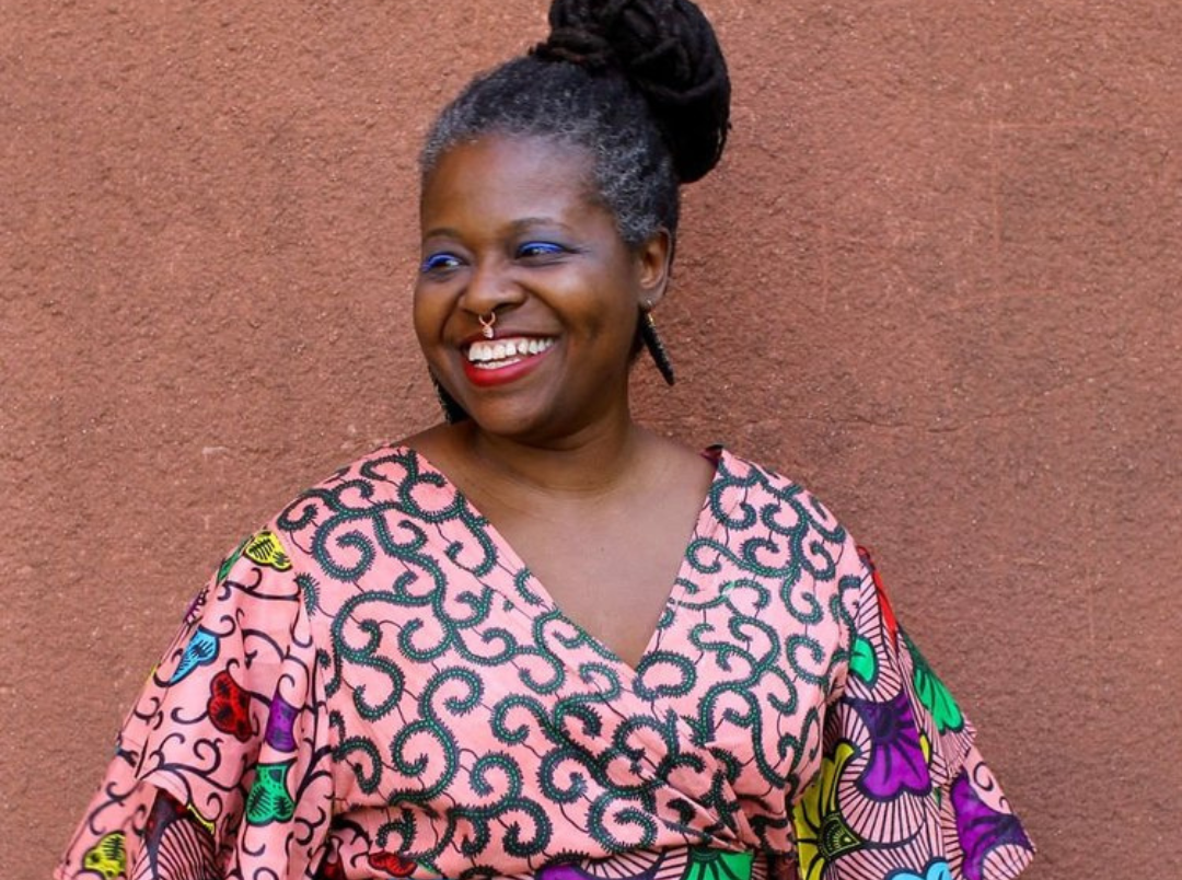 Smiling person with a colorful patterned outfit stands against a textured brown wall. Their hair is styled in a bun, and they wear large earrings.