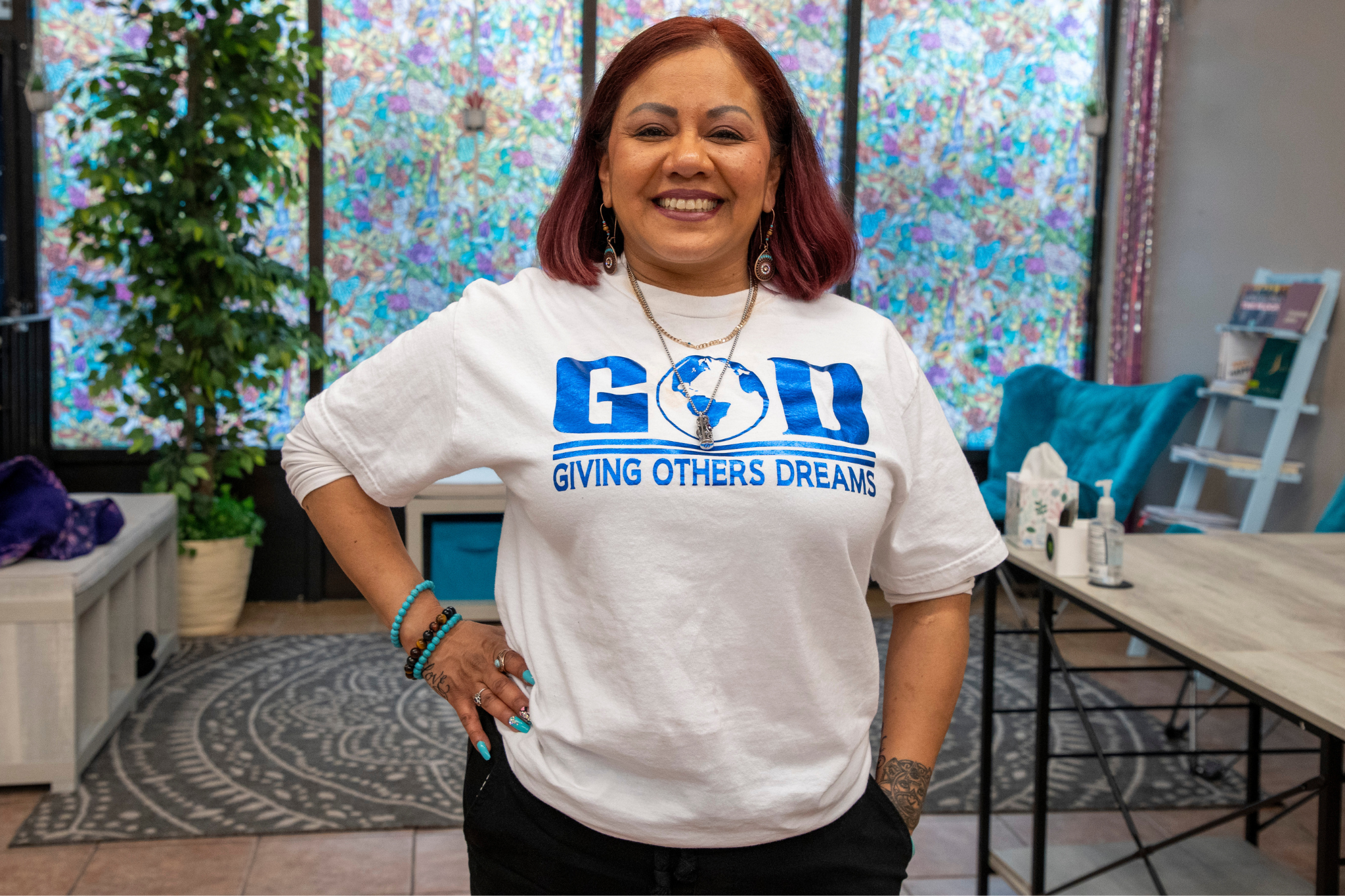 Smiling woman with red hair stands in a room with colorful decor. She wears a white shirt with blue text GOD Giving Others Dreams and has one hand on her hip. A plant and bright artwork are in the background.