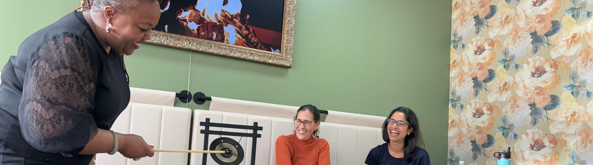 Two women smiling while looking at a women wearing a black long sleeve shirt rings a miniature gong. The background is a green wall with an accent wall that has a pattern of yellow and white flowers.
