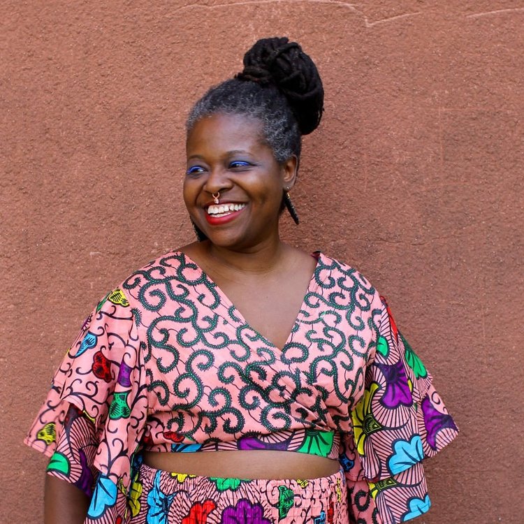 Smiling person with a colorful patterned outfit stands against a textured brown wall. Their hair is styled in a bun, and they wear large earrings.