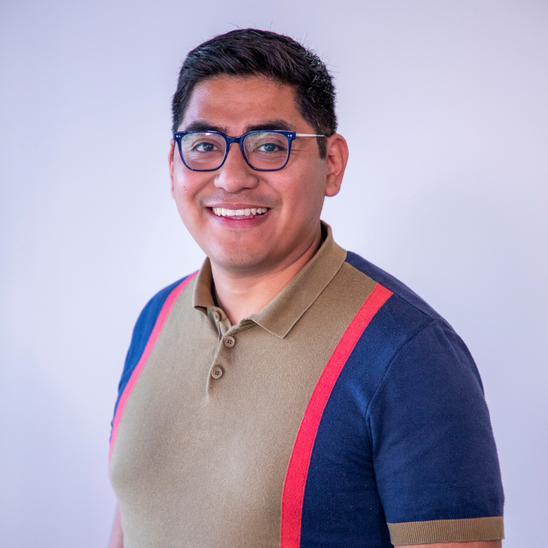 Smiling person with short dark hair and glasses wearing a collared shirt with colorful stripes, standing against a plain background.