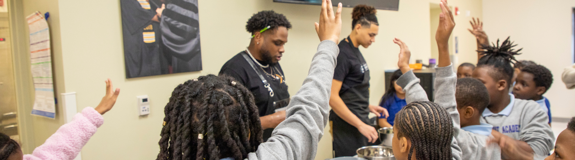 A man with a cooking apron teaching small children how to cook. The children have their hands raised.