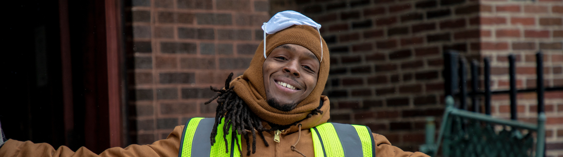 A person wearing a brown coat, scarf, and green safety vest stands with arms outstretched, smiling. They have long hair, and a blue face mask is on top of their head. A brick wall and iron fence are in the background.