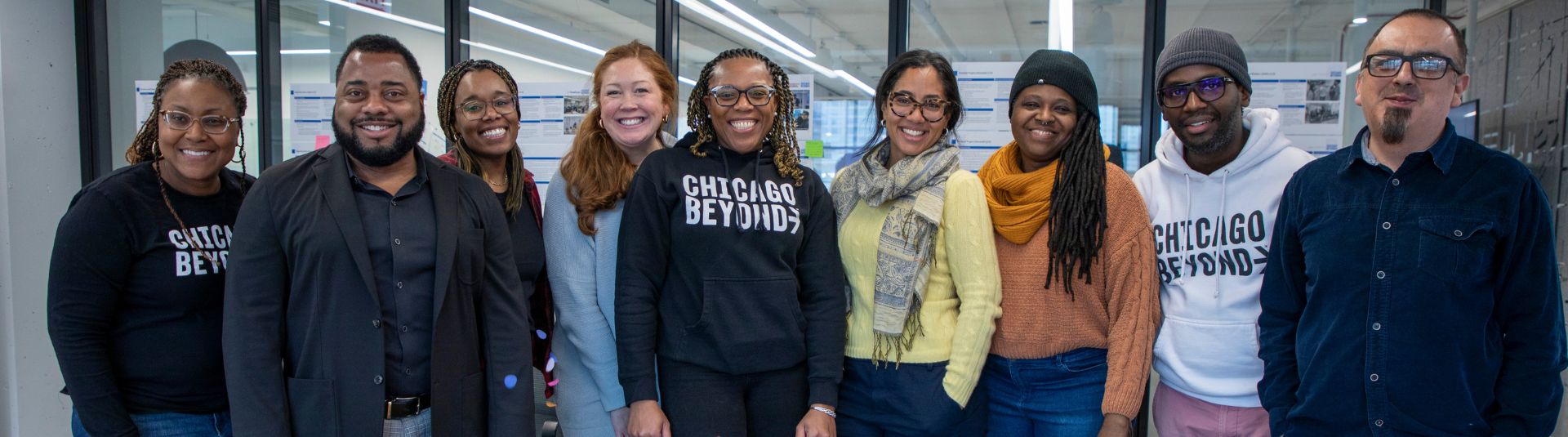 A group of nine people pose together indoors, some wearing "Chicago Beyond" shirts, standing in front of glass wall panels.