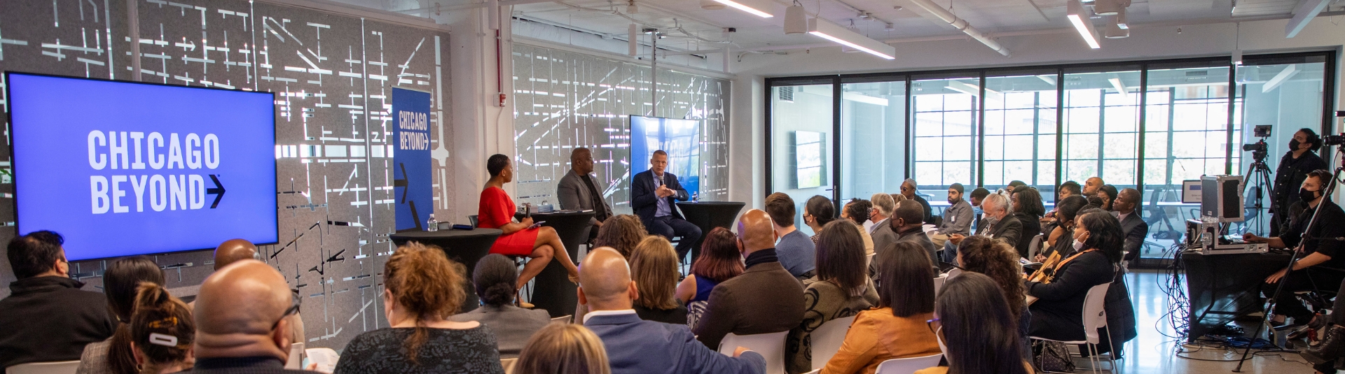 A large room where filled with people sitting down watching a panel conversation of three people talking. Two televisions have Chicago Beyond logo on their screen.