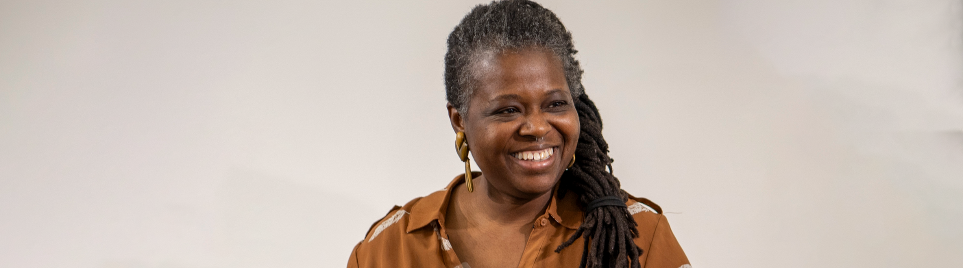 A person with long dreadlocks smiles while wearing a brown shirt. The background is plain and softly blurred.